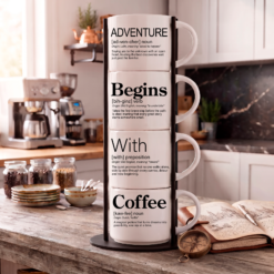 Adventure Begins With Coffee mug set, four 14-ounce white stackable mugs in Word Story Style in a black metal Storyteller Display Stand on a rustic coffee bar with a compass and travel journal.