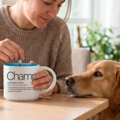Personalized pet name mug reading Champ in Whimsical Word Story Style with Ocean Blue interior, woman stirring coffee as a golden retriever rests his head on the table.