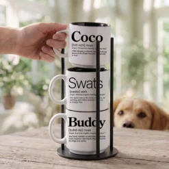 Personalized pet name mug set reading Coco Swats Buddy, three 14-ounce white stackable mugs with black interiors in a black metal Storyteller Display Stand, golden retriever peeking over the table.