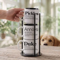 Personalized pet name mug set reading Pickles Annoys Duke, three 14-ounce white stackable mugs with black interiors in a black metal Storyteller Display Stand, golden retriever peeking over the table