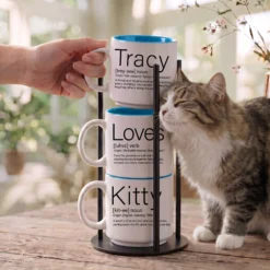 Personalized pet parent name mug set reading Tracy Loves Kitty, three 14-ounce white stackable mugs with Ocean Blue interiors in a black metal Storyteller Display Stand, tabby cat rubbing against the stack.