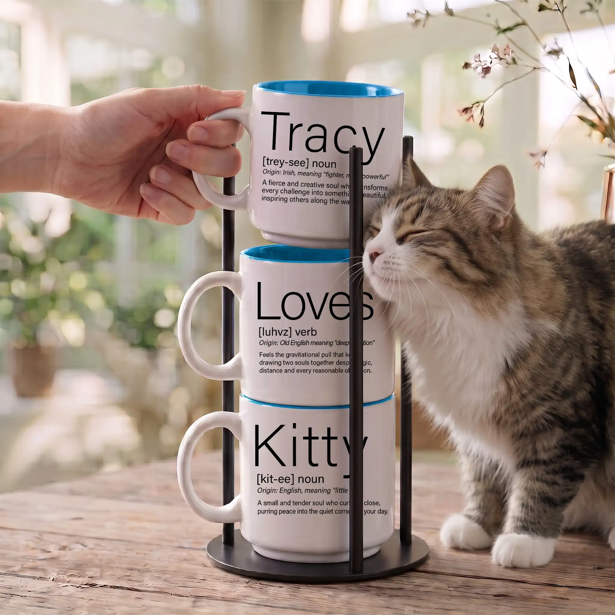 Personalized pet parent name mug set reading Tracy Loves Kitty, three 14-ounce white stackable mugs with Ocean Blue interiors in a black metal Storyteller Display Stand, tabby cat rubbing against the stack.