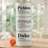 Personalized pet name mug set reading Pickles Annoys Duke, three 14-ounce white stackable mugs with black interiors in a black metal Storyteller Display Stand, golden retriever peeking over the table.